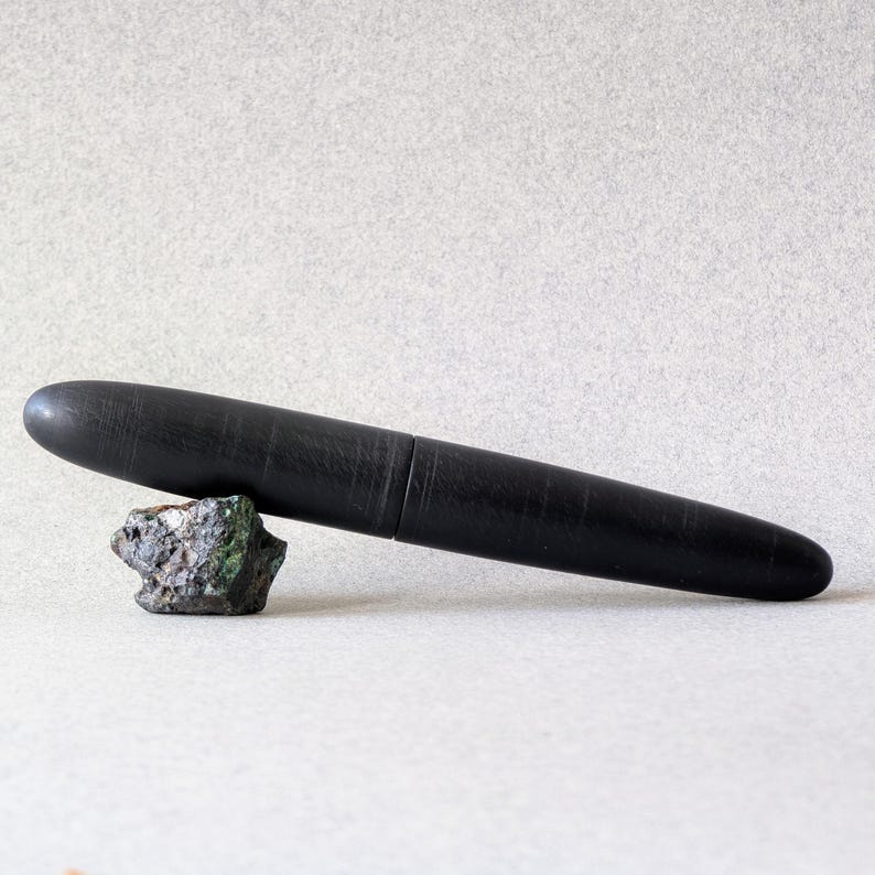 Peut inclure: Un stylo noir &agrave; la surface textur&eacute;e repose sur une petite roche multicolore. Le stylo a un design &eacute;l&eacute;gant et cylindrique avec un capuchon. Le fond est de couleur gris clair uni.