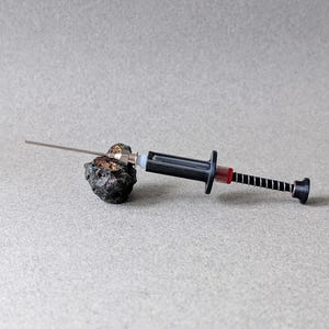 Könnte beinhalten: Eine schwarze und silberne Spritze mit einer langen Nadel liegt auf einem dunklen, strukturierten Stein. Die Spritze hat einen schwarzen Kolben und einen roten Akzent in der Nähe der Nadel. Der Hintergrund ist einfarbig hellgrau.