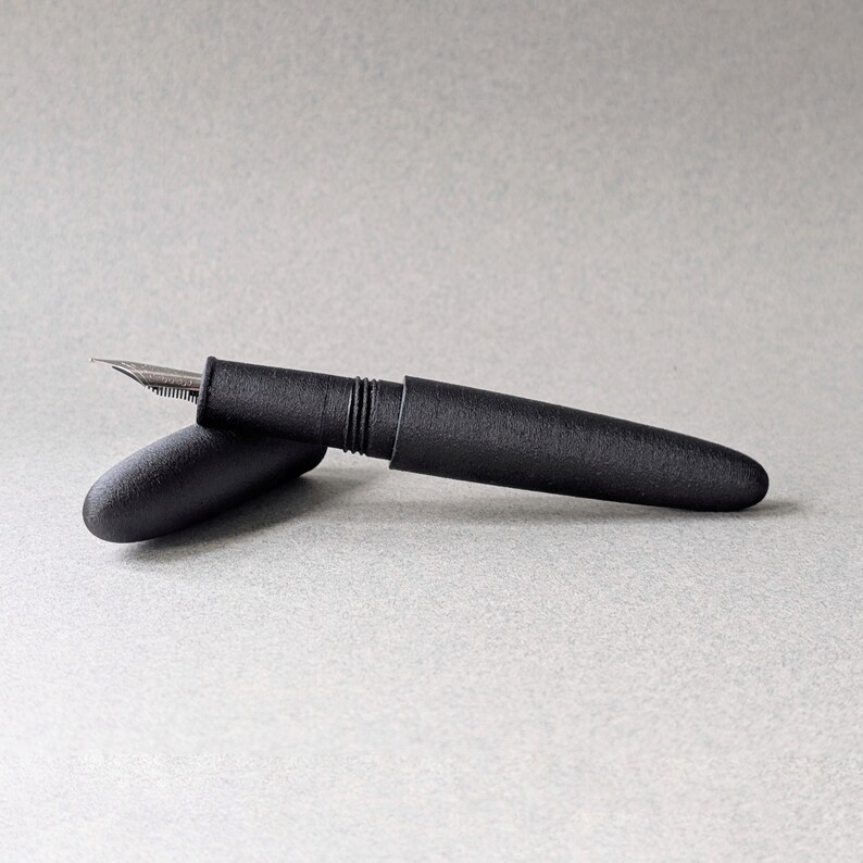 Peut inclure: Un stylo plume noir mat avec une plume argent&eacute;e et un capuchon. Le stylo est pos&eacute; sur une surface gris clair. Le capuchon est ouvert, r&eacute;v&eacute;lant la plume. Le stylo a un design &eacute;pur&eacute; et minimaliste.