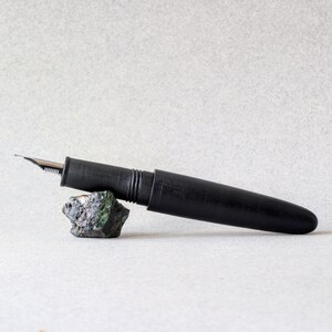 Peut inclure: Un stylo plume noir avec une plume argent&eacute;e repose sur une petite roche textur&eacute;e aux teintes vertes et grises. Le stylo est cylindrique avec un capuchon et une finition lisse et mate. Le fond est de couleur claire et neutre.