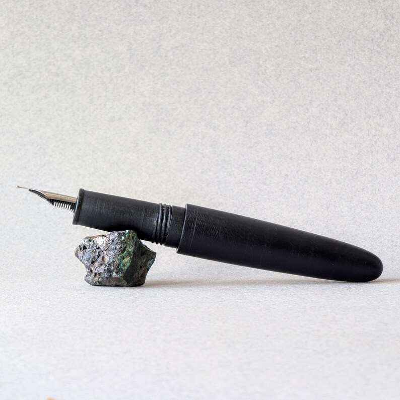 Peut inclure: Un stylo plume noir avec une plume argent&eacute;e repose sur une petite roche textur&eacute;e aux teintes vertes et grises. Le stylo est cylindrique avec un capuchon et une finition lisse et mate. Le fond est de couleur claire et neutre.