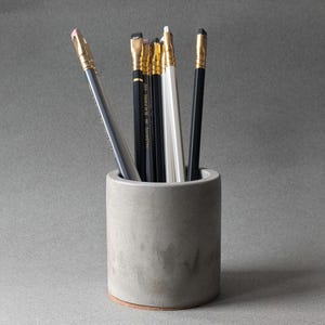 May include: A gray concrete pencil holder with six pencils inside. The pencils are black, white, and gray with gold accents.