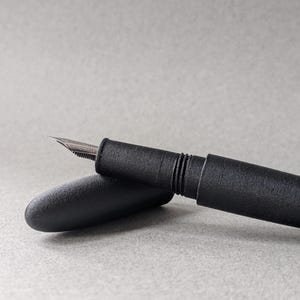 Peut inclure: Un stylo plume noir avec une plume argent&eacute;e et un capuchon. Le stylo est pos&eacute; sur une surface grise. Le corps du stylo a une finition mate.