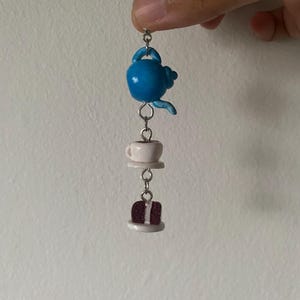 Peut inclure: Un charm fantaisiste représentant une théière bleue, une tasse à thé sur une soucoupe et une part de gâteau. Les breloques sont reliées par des anneaux argentés, créant un thème de mini tea party. La théière est bleu vif et le gâteau a une garniture blanche.
