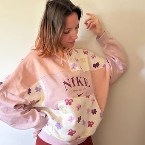 Könnte beinhalten: Ein farblich abgesetzter Kapuzenpullover in Hellrosa und Creme mit einem wiederkehrenden Muster aus kleinen Schmetterlingen in Lila, Pink und Gelb. Auf der Brust steht der Text NIKE und Athletic dept. 1972 in Dunkellila mit einem kleinen Swoosh-Logo.