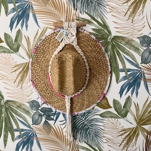 Op de afbeelding: Een witte macramé wandhanger met een kwast, hangend aan een houten stok, met een strohoed met een roze en witte bloemenrand eronder.