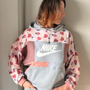 Puede incluir: Una sudadera con capucha de bloques de color que presenta una tela rosa claro con un patrón floral rojo y azul en las mangas y la parte superior del pecho, combinada con paneles texturizados grises y rosa empolvado. Incluye el logo Nike en blanco.