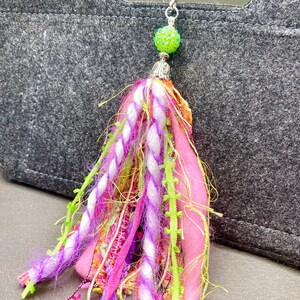 May include: A colorful tassel keychain with a silver clasp. The tassel features pink, purple, white, and green threads, with a green beaded ball at the top. The keychain is attached to a gray felt bag.