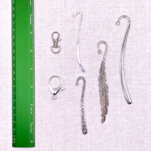 May include: An assortment of metal bookmarks and clasps. The bookmarks include a silver feather design, a curved silver bookmark, and a decorative bookmark. A green ruler with inch measurements is also present.