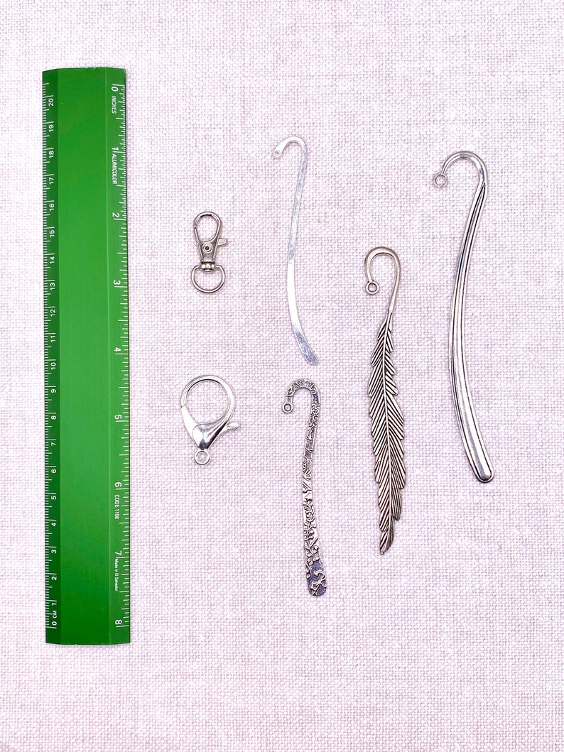 May include: An assortment of metal bookmarks and clasps. The bookmarks include a silver feather design, a curved silver bookmark, and a decorative bookmark. A green ruler with inch measurements is also present.