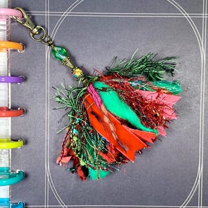 May include: A colorful tassel charm with a brass clasp and green bead. The tassel features red, green, and pink fabric strips and strands, creating a vibrant and textured accessory. The charm is attached to a binder.