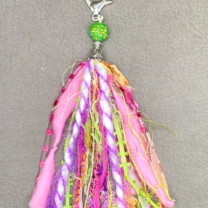 May include: A colorful tassel keychain with a silver clasp and a green, glittery bead. The tassel features pink, purple, white, and green fabric strands, creating a vibrant and textured accessory.