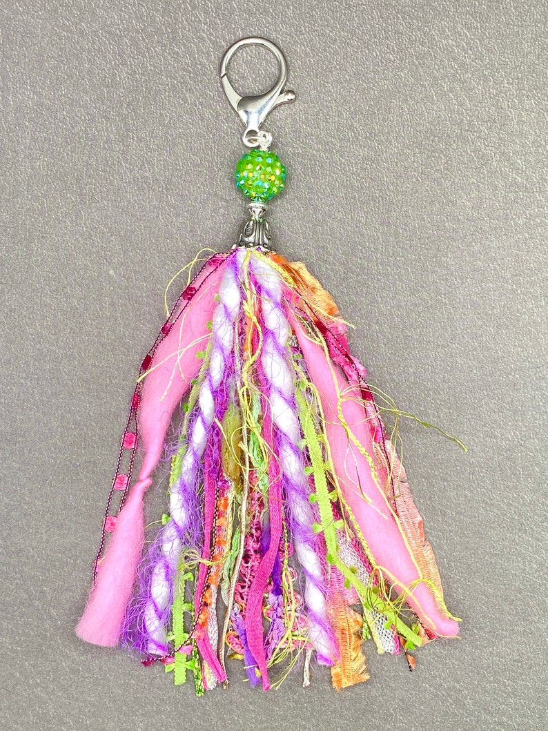 May include: A colorful tassel keychain with a silver clasp and a green, glittery bead. The tassel features pink, purple, white, and green fabric strands, creating a vibrant and textured accessory.