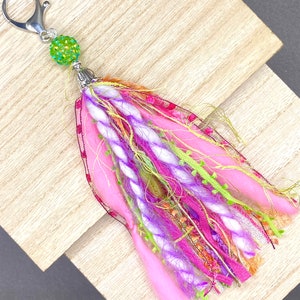 May include: A handmade keychain tassel with a silver clasp and a green beaded accent. The tassel features pink, purple, and green fabric and yarn, creating a colorful and textured accessory. This item is perfect for adding a unique touch to keys or bags.