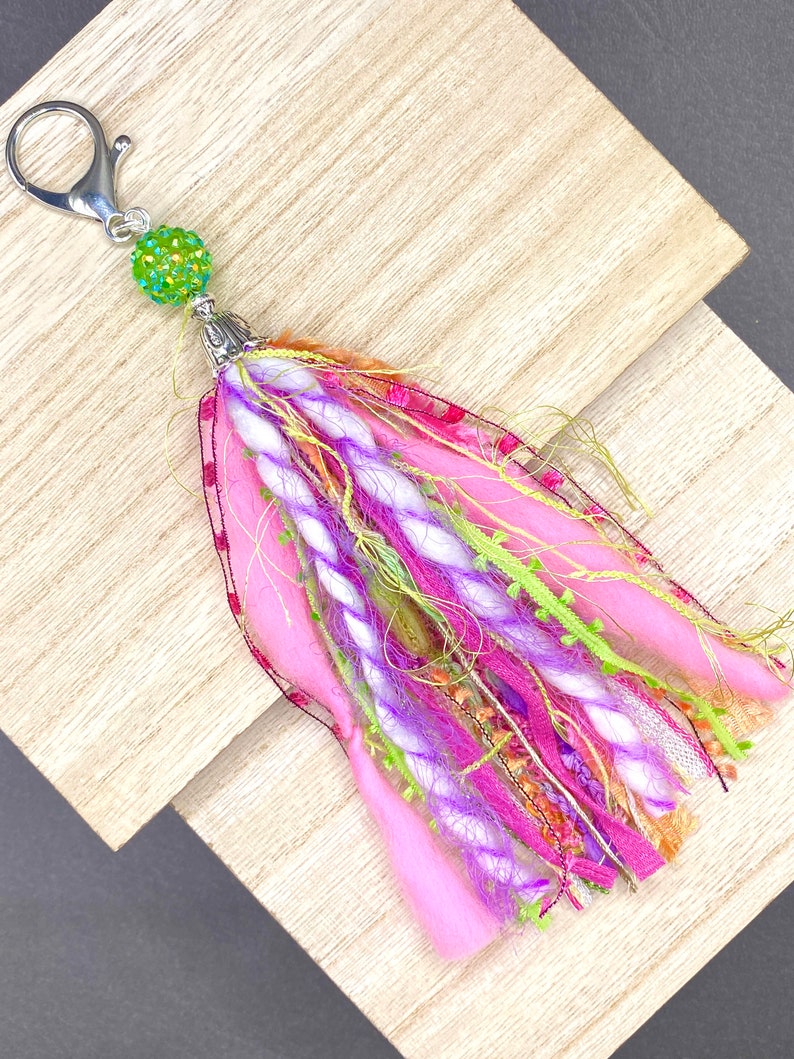 May include: A handmade keychain tassel with a silver clasp and a green beaded accent. The tassel features pink, purple, and green fabric and yarn, creating a colorful and textured accessory. This item is perfect for adding a unique touch to keys or bags.