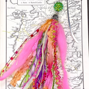 May include: A handmade bookmark with a silver metal design and a colorful tassel. The tassel features pink, orange, purple, and green ribbons, yarn, and beads. The bookmark is on a map of a fictional land with the text "The South".