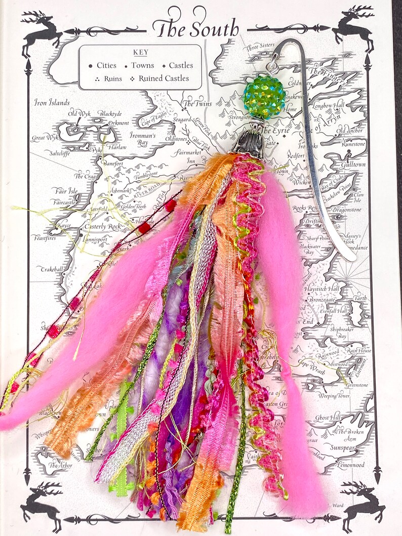 May include: A handmade bookmark with a silver metal design and a colorful tassel. The tassel features pink, orange, purple, and green ribbons, yarn, and beads. The bookmark is on a map of a fictional land with the text "The South".