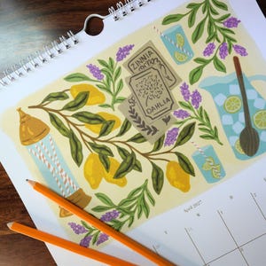 May include: A calendar page with a botanical illustration featuring lemons, lavender, and seed packets labeled "Zinnia" and "Dahlia." The artwork includes a pitcher of lemonade, two glasses, and a wind chime. Two orange pencils rest on the page.