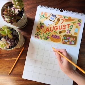 May include: A wall calendar open to August, with a vibrant illustration. The artwork includes travel motifs such as a camera, suitcase, and hat. Two potted succulents and a pencil are on a wooden surface. The calendar is being written on.