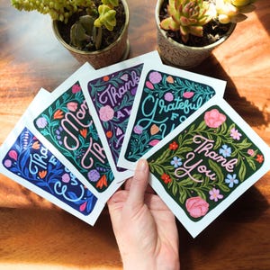 May include: A set of four rectangular greeting cards featuring floral illustrations and hand-lettered text. The cards display phrases including Thank You and Grateful For You, surrounded by colorful botanical patterns in pink, blue, and green.