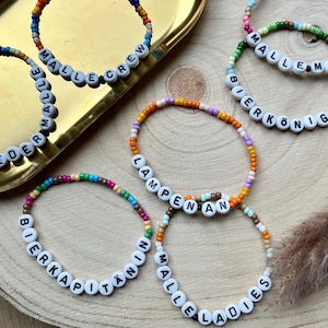 May include: Several beaded bracelets with white letter beads spelling out phrases such as "MALLE CREW" and "ENDLICH WIEDER MALLE". The bracelets have colourful beads and are displayed on a gold tray and a wooden surface.