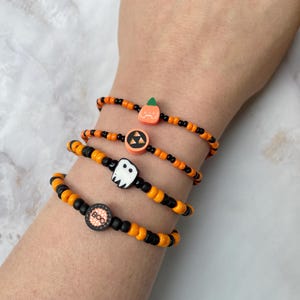 Op de afbeelding: Vier kralenarmbanden met afwisselend oranje en zwarte rocailles. Elke armband bevat een unieke bedel: een oranje pompoen, een zwart-oranje cirkelvormig triforce-symbool, een wit spookje en een zwarte kraal met de tekst BOO.