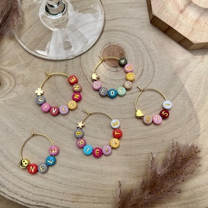 Op de afbeelding: Vijf goudkleurige oorringen met kleurrijke ronde kralen met letters die de namen Vivienne, Geoffrey, Susi, Brigitte en Nele spellen. Elke ring bevat kleine goudkleurige bedels in de vorm van sterren, hartjes, kronen of smileygezichtjes.