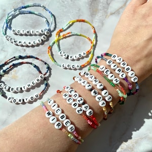 Könnte beinhalten: Armbänder mit weißen Buchstabenperlen, die Sätze wie "NOTHING COMPARES" und "I'LL BE THERE FOR YOU" bilden. Die Armbänder haben bunte Perlen und werden am Handgelenk getragen. Weitere Sätze sind "LA VIDA LOCA".
