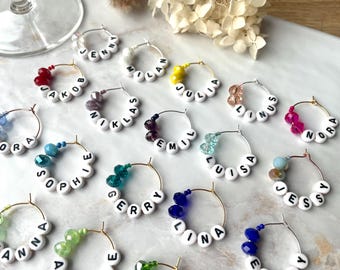 May include: A set of 14 gold-toned wine glass charms with different colored beads and white beads spelling out names like "Jenny", "Milan", "Julia", "Linus", "Nora", "Zora", "Jakob", "Niklas", "Emil", "Louisa", "Jessy", "Gerry", "Lina", and "Hanna".