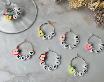 May include: Six gold and silver wine glass charms with apple charms and names spelled out in white beads. The names are Ida, Mia, Emilia, Nora, Leonie, and Lina.