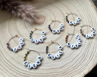 May include: Seven circular hoop earrings made of thin gold-toned metal wire. Each hoop features a segment of small, colorful glass seed beads and white plastic alphabet beads spelling out the names Juli, Livia, Moni, Alex, Liam, Niklas, and Elena.