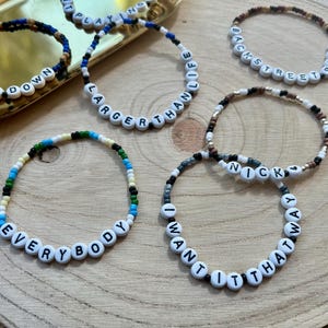 Può includere: Una collezione di braccialetti di perline con perline di lettere bianche che compongono frasi come "GET DOWN", "LARGER THAN LIFE", "BACKSTREET BOYS", "EVERYBODY" e "I WANT IT THAT WAY". I braccialetti presentano vari schemi di colori.
