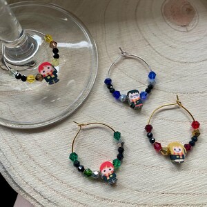 Op de afbeelding: Set van vier wijnglasbedels. Elke bedel heeft een klein karakterfiguur en kleurrijke kralen. De bedels zitten aan zilveren en gouden ringen. Op een houten oppervlak.