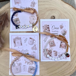 Könnte beinhalten: Drei Perlenarmbänder auf Präsentationskarten. Ein Armband hat blaue und goldene Perlen mit dem Namen VRENI und einem goldenen Fuß-Anhänger. Ein weiteres hat hellblaue und beige Perlen mit dem Namen MONA und einem Elefanten-Anhänger. Das dritte hat bernsteinfarbene Perlen mit dem Wort BABYBOY und einem Marienkäfer-Anhänger.