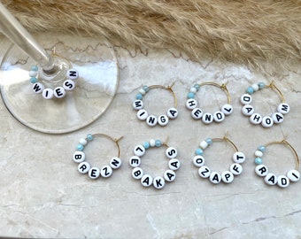 May include: Gold-toned wine charms with white letter beads spelling out various names. Each charm features a ring of white beads with black letters, accented by light blue and white beads. A wine glass is in the image.