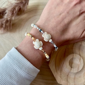 Könnte beinhalten: Zwei Armbänder mit Schildkrötenanhängern. Ein Armband hat einen weißen Schildkrötenanhänger mit weißen und silbernen Perlen. Das andere Armband hat einen weißen Schildkrötenanhänger mit gelben und braunen Perlen.