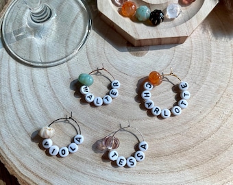May include: Four personalized wine charms with names spelled out in white letter beads. Each charm has a different colored bead accent. The charms are on a wooden surface with a wine glass in the background.