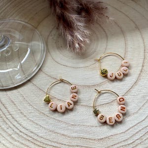 Può includere: Tre orecchini a cerchio dorati con perline rosa che compongono nomi e una faccina sorridente. Gli orecchini sono su una superficie di legno con un bicchiere da vino e fiori secchi sullo sfondo.