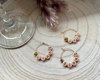 May include: Three gold hoop earrings with pink beads spelling out names and a smiley face. The earrings are on a wooden surface with a wine glass and dried flowers in the background.