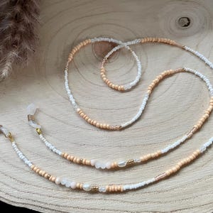 Op de afbeelding: Een brilketting gemaakt van witte en perzikkleurige kralen. De ketting heeft twee goudkleurige sluitingen om aan de bril te bevestigen.