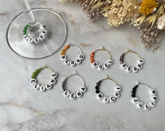 May include: A collection of circular metal wine glass charms featuring white alphabet beads spelling the names STEFFI, BERND, NOEL, GERRY, ELIANE, LENA, MICHI, and JULIAN. Each charm is accented with small, colored faceted beads in green, orange, red, purple, grey, and black.