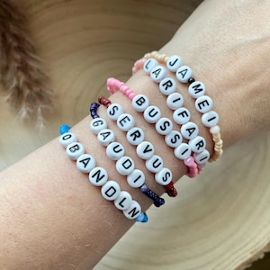May include: A collection of beaded bracelets spelling out names and words. The bracelets are made with white letter beads and colorful string. The names and words include "OBANDLN", "GAUDI", "SERVUS", "BUSSI", "LARI", and "JAMEI".