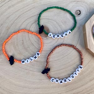 Op de afbeelding: Drie kralenarmbanden met kleine ronde kralen in oranje, groen en metallic koper. Elke armband bevat witte letterkralen die de namen Wilma, Verena en Charlotte spellen, samen met een kleine zwarte bedel in de vorm van een vleermuis.