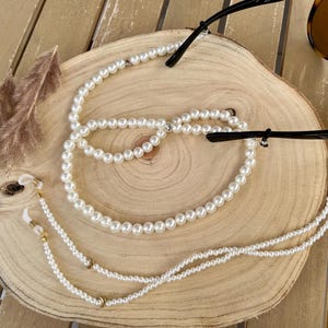 Op de afbeelding: Een brilketting van witte parels met kleine, ronde, glanzende kralen. De ketting is voorzien van goudkleurige metalen accenten en doorzichtige siliconen lusjes aan beide uiteinden voor bevestiging aan de pootjes van een bruine schildpadzonnebril.