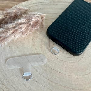 May include: A black phone case with a textured pattern, a clear phone strap, and a silver ring. The phone case is placed on a light brown wooden surface, with dried ornamental grass in the background.