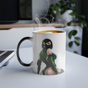 May include: A white ceramic mug with a black handle. The mug has a color-changing design that reveals a woman in a green and black outfit when hot liquid is added. The woman is wearing a black mask and is looking over her shoulder.