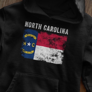 May include: Black hoodie with a distressed North Carolina flag graphic. The flag features the state seal with the text "May 20th 1775" and "April 12th 1776".