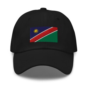Op de afbeelding: Een zwarte baseballpet met een geborduurde vlag van Namibië. De vlag heeft een rode diagonale streep, een blauwe driehoek met een gele zon en een groen ondergedeelte.