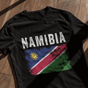 Peut inclure: T-shirt noir avec un graphique délavé blanc du drapeau namibien et le mot "NAMIBIA" au-dessus.