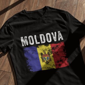 Könnte beinhalten: Schwarzes T-Shirt mit einem abgenutzten weißen Druck des Wortes "MOLDOVA" und der moldauischen Flagge.